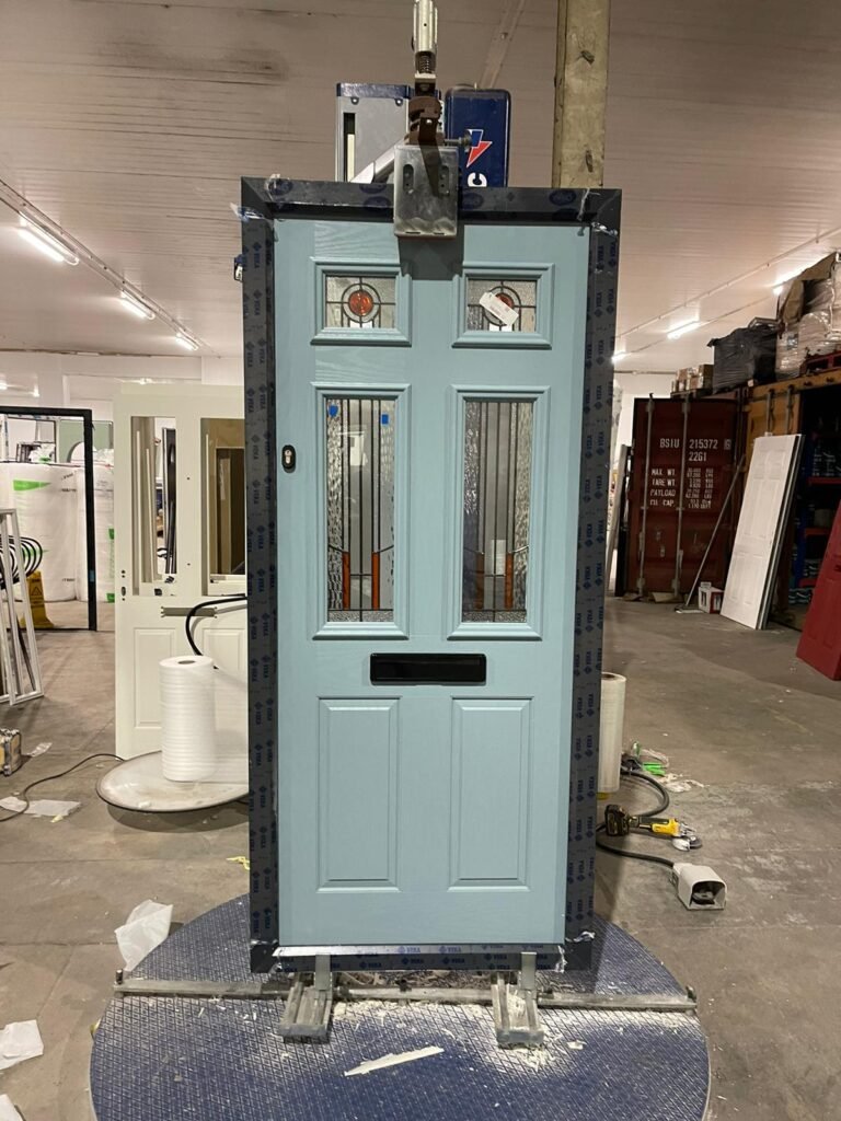 Pale blue front door with decorative leaded glass panels and a black letterplate, being wrapped on a turntable in the factory